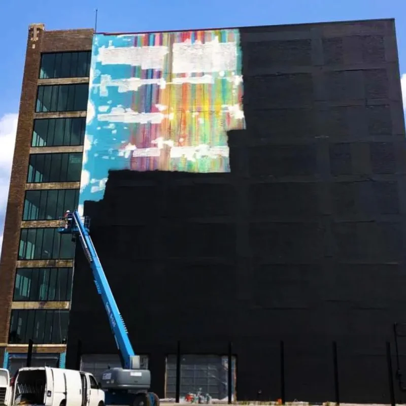 High-rise building wall with the old mural partly covered in black base coat, boom lift working the upper section