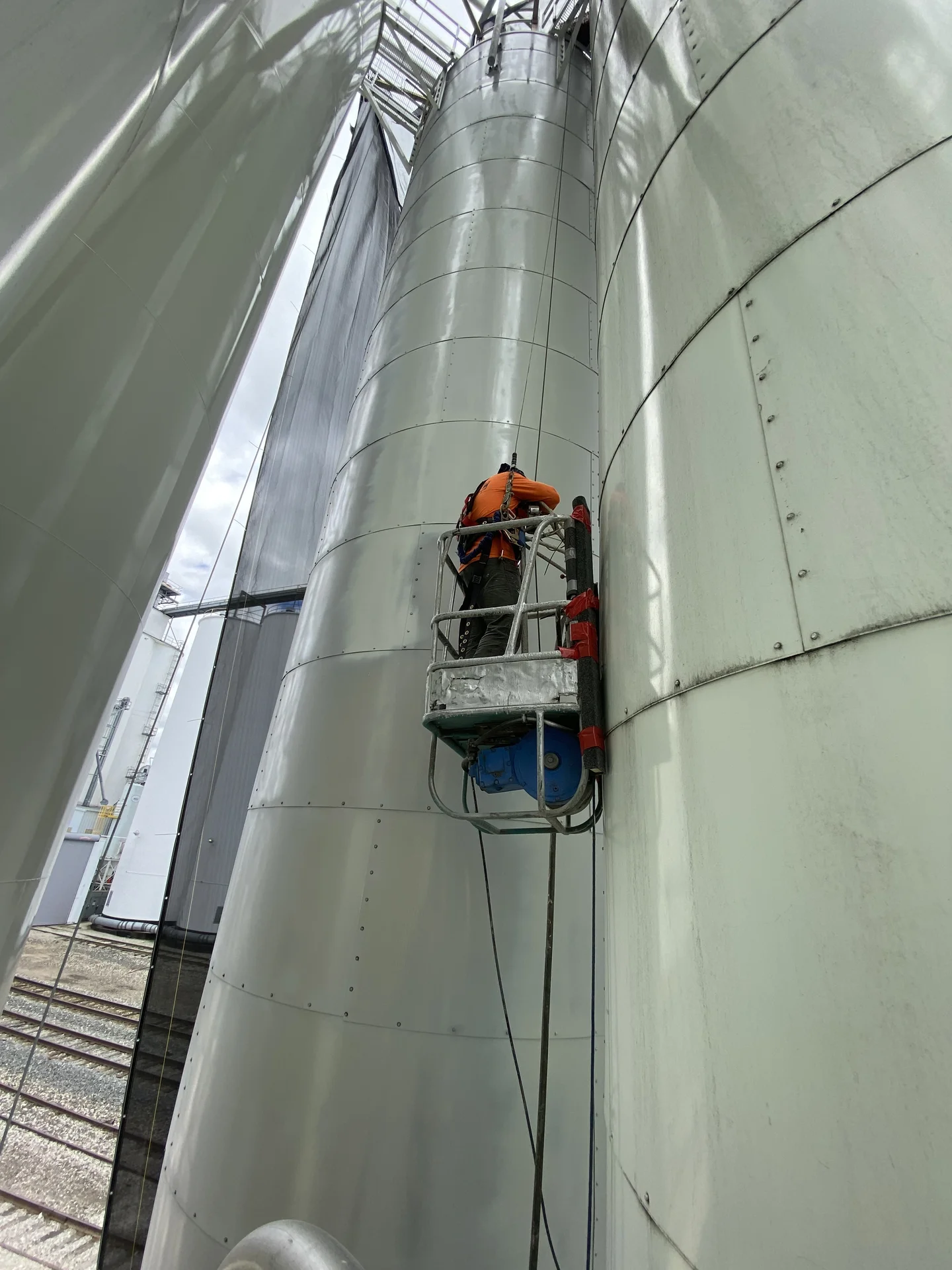 Two crew members on a suspended platform painting the exterior of a large silo at height