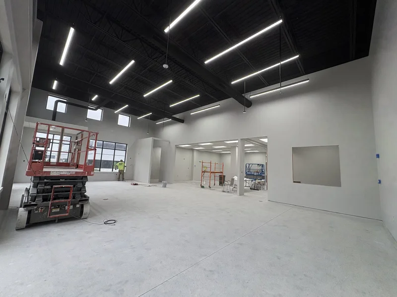 Wide view of the Pella interior buildout — black ceiling, steel beams, and fresh gray walls with lifts on site during painting